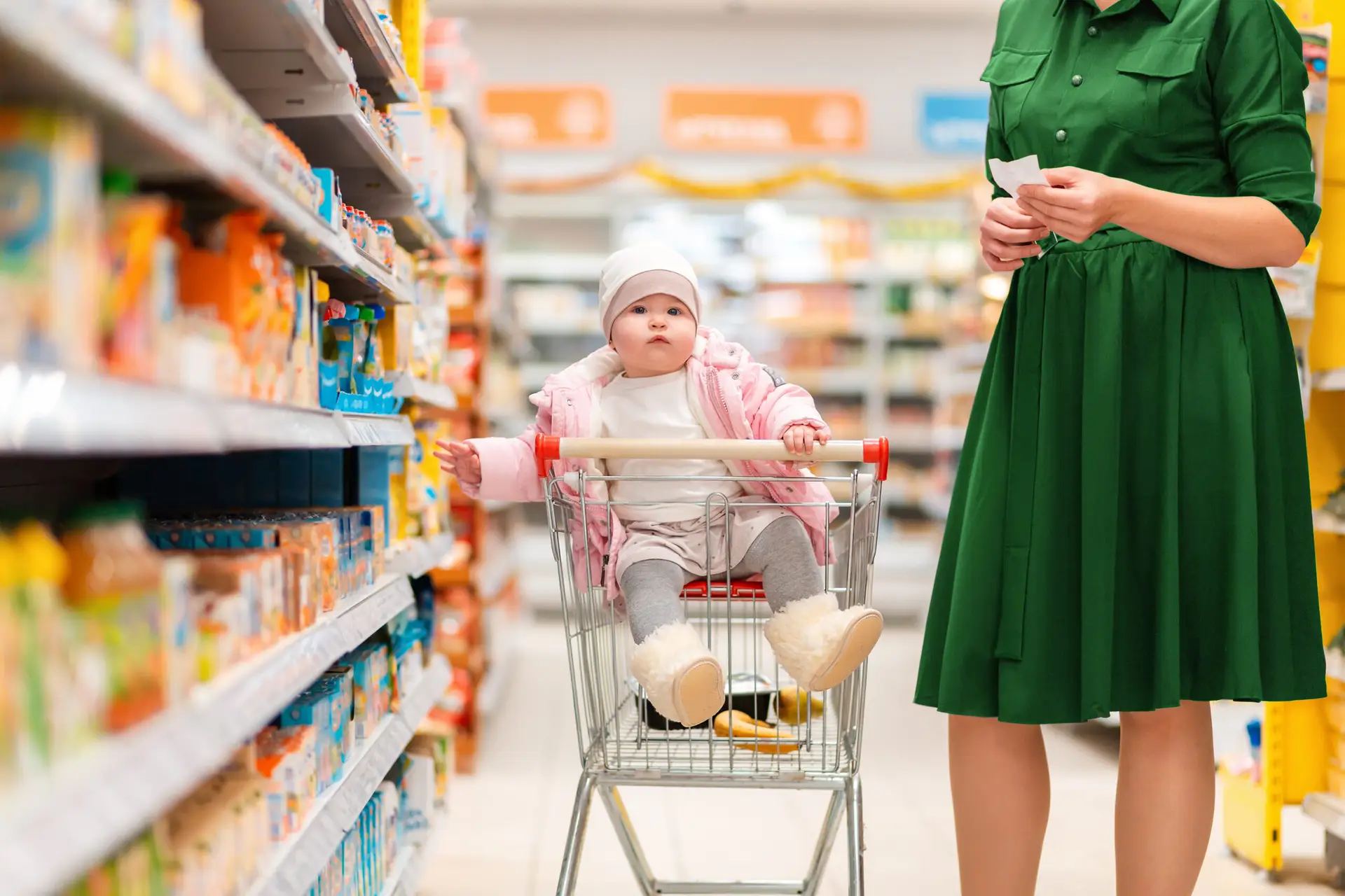 A young mother and her baby sitting in a grocery cart choose food on a supermarket shelf.